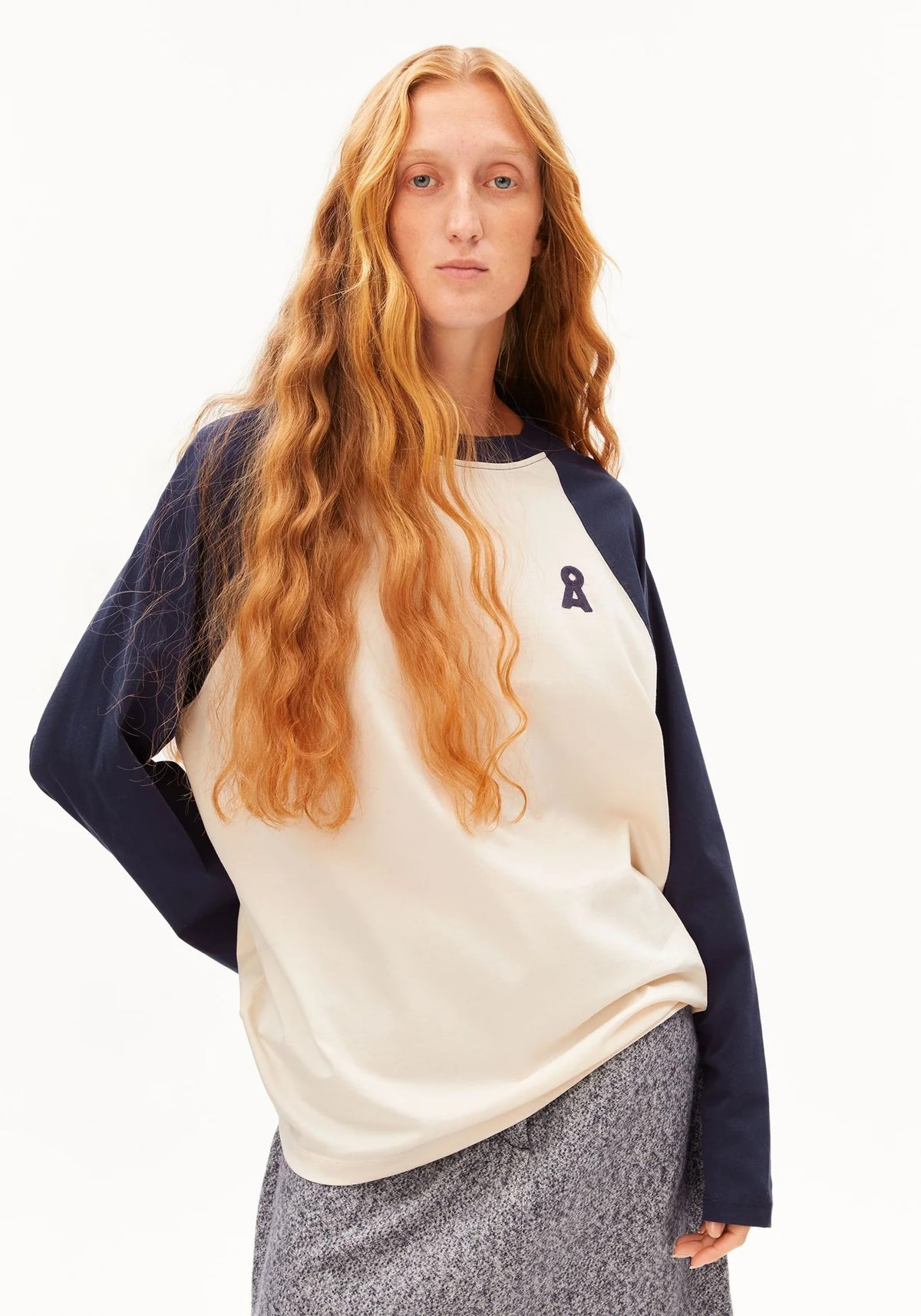 A woman with long wavy red hair wearing a cream and navy raglan sweatshirt with a small navy ribbon logo on the left chest, paired with a gray textured skirt, against a plain white studio background.