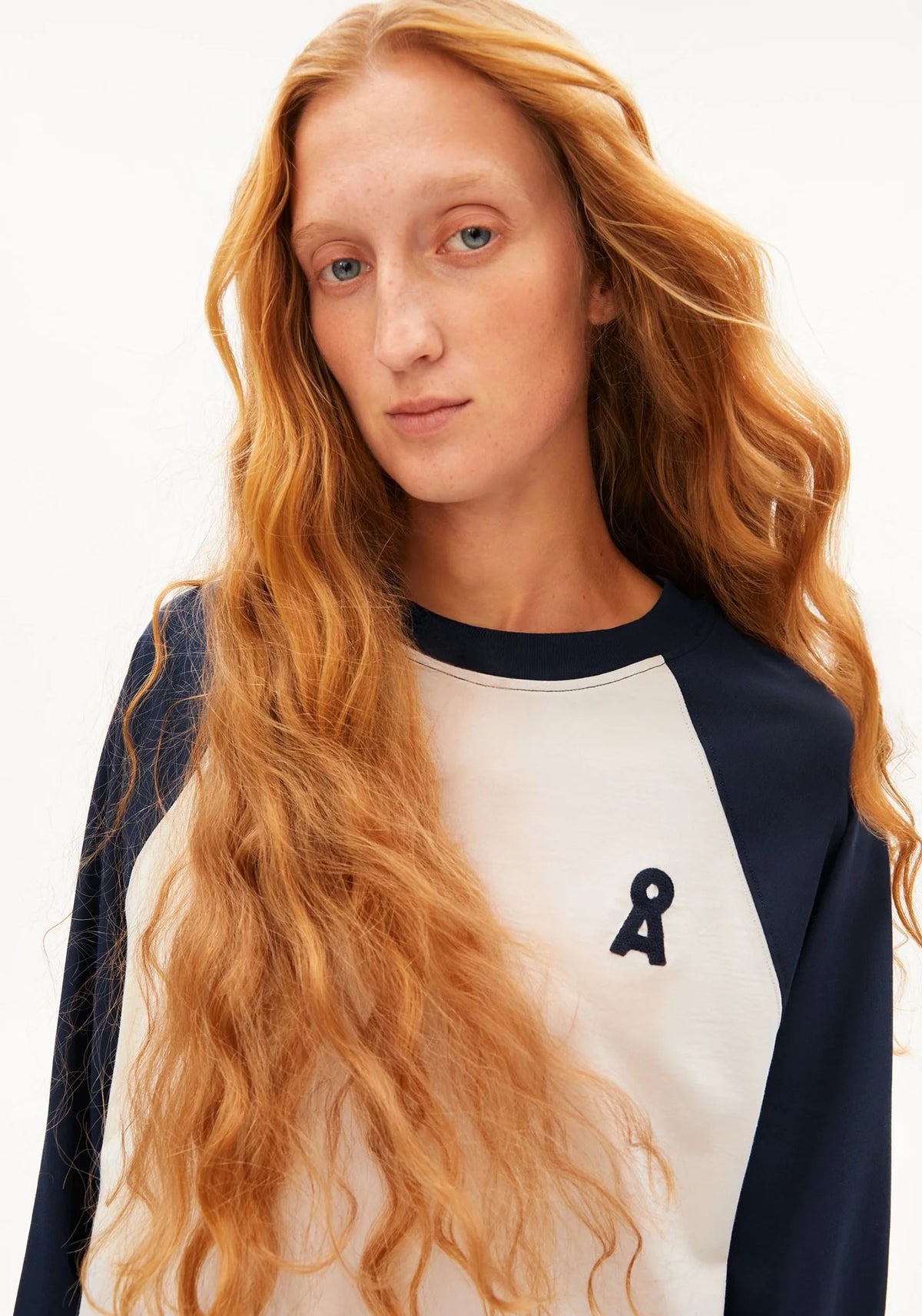 A woman with long wavy red hair wearing a navy and off-white raglan sweatshirt with a small navy graphic on the chest, photographed against a clean white background.