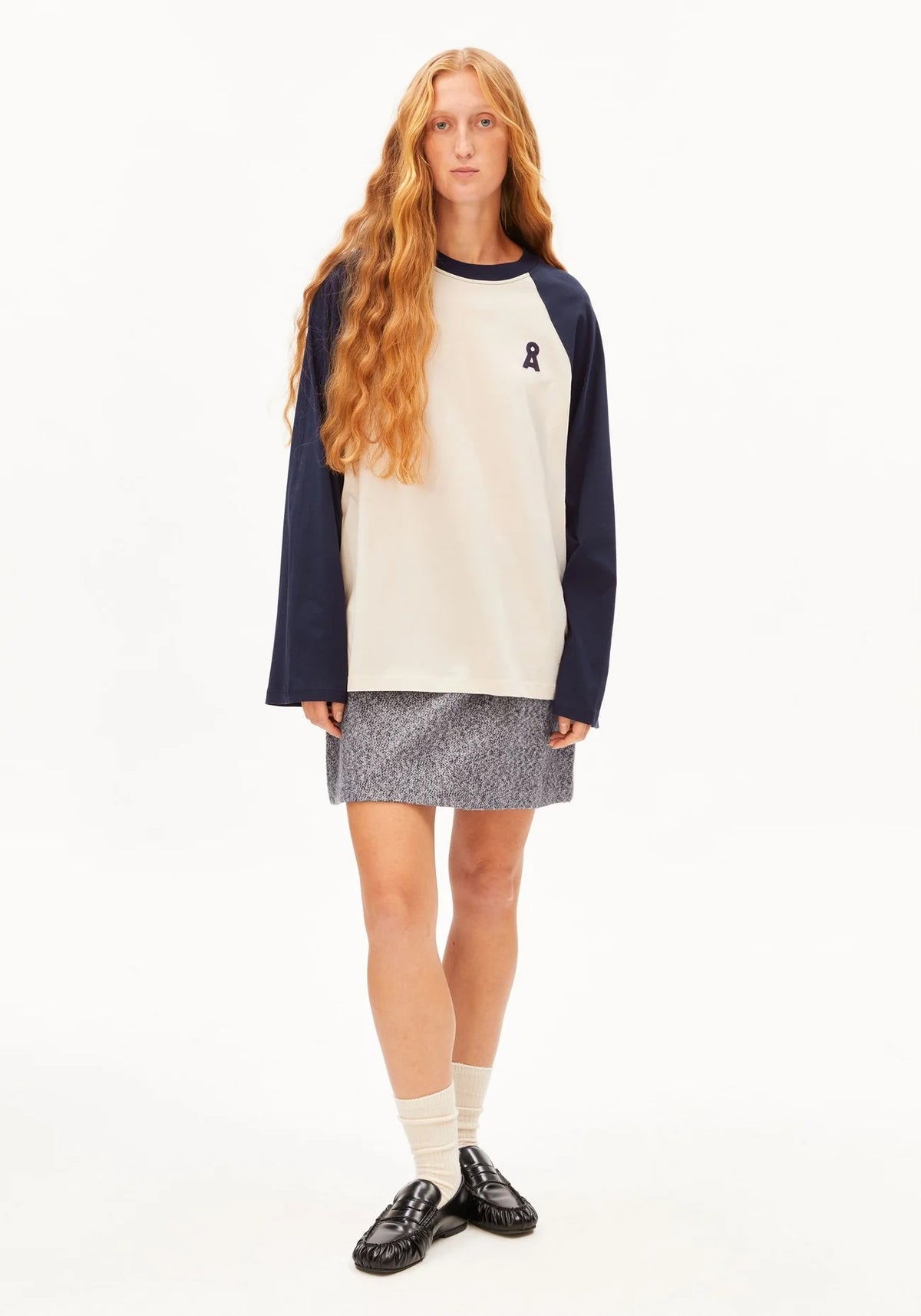 A model with long red hair stands against a white studio background wearing a colorblock raglan long-sleeve shirt with navy sleeves and collar, an off-white body, a gray patterned mini skirt, cream socks, and black leather loafers.