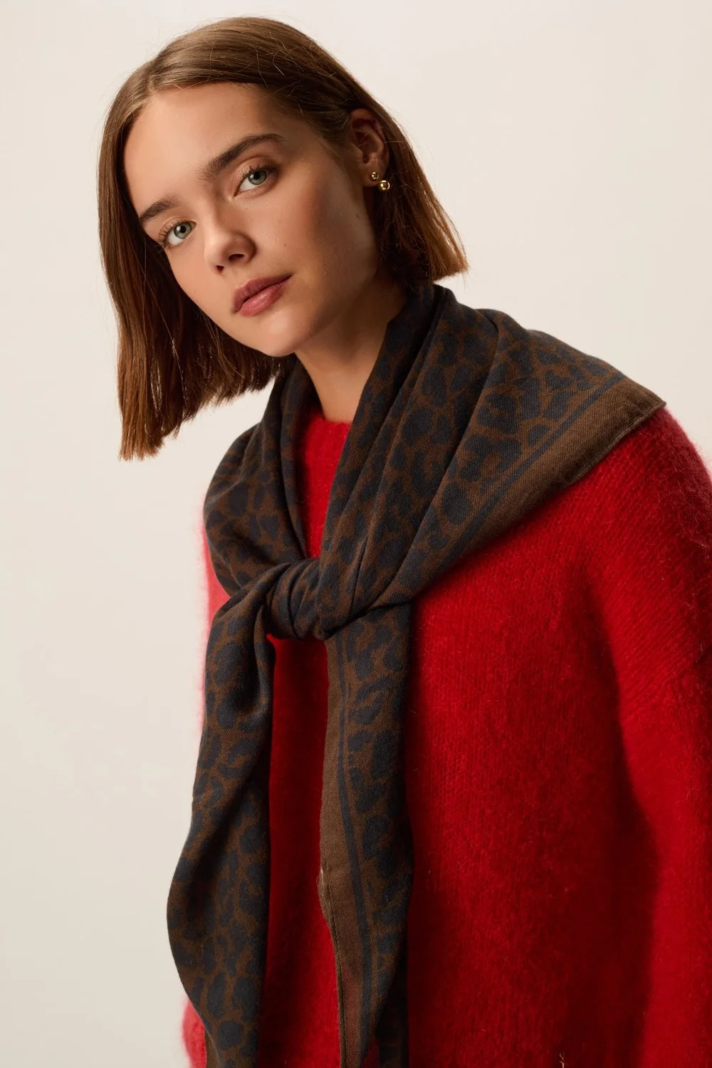 A woman with short brown hair models a dark brown leopard-print scarf wrapped around her neck over a bright red knit sweater, set against a plain off-white background.