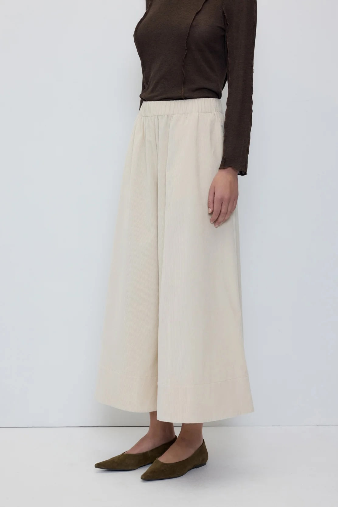 A model is shown from the chest down wearing a dark brown long-sleeve top, high-waisted off-white wide-leg cropped pants, and olive green flats, set against a plain white studio background.