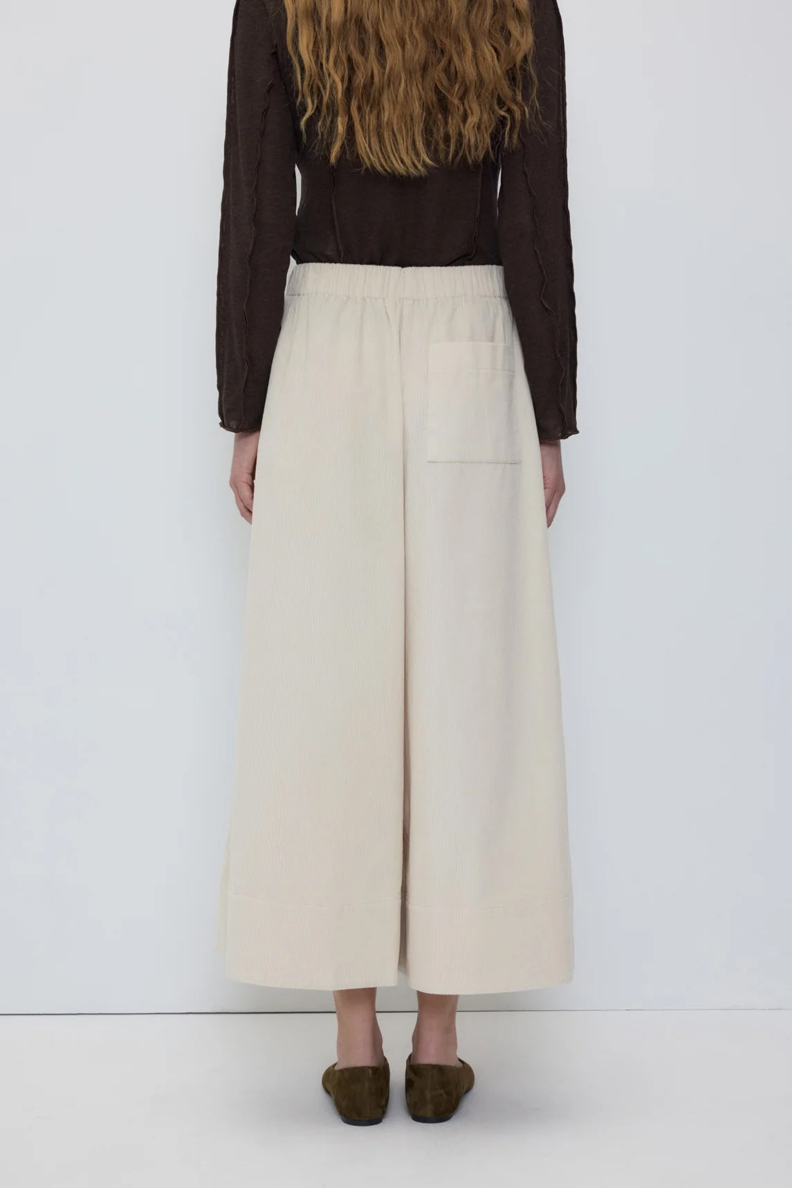 A model shown from the back wearing a dark brown long-sleeve top and high-waisted, wide-leg cream trousers with a side pocket, paired with dark brown shoes.