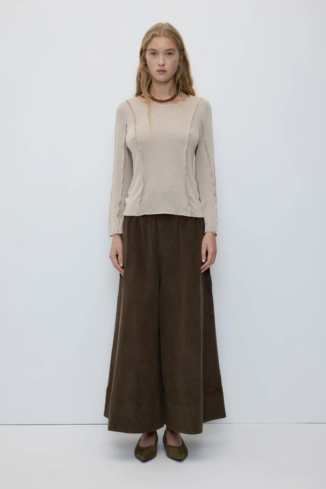 A woman stands against a plain light gray studio background wearing a beige long-sleeve knit top and wide-leg dark brown trousers, with brown pointed flats.