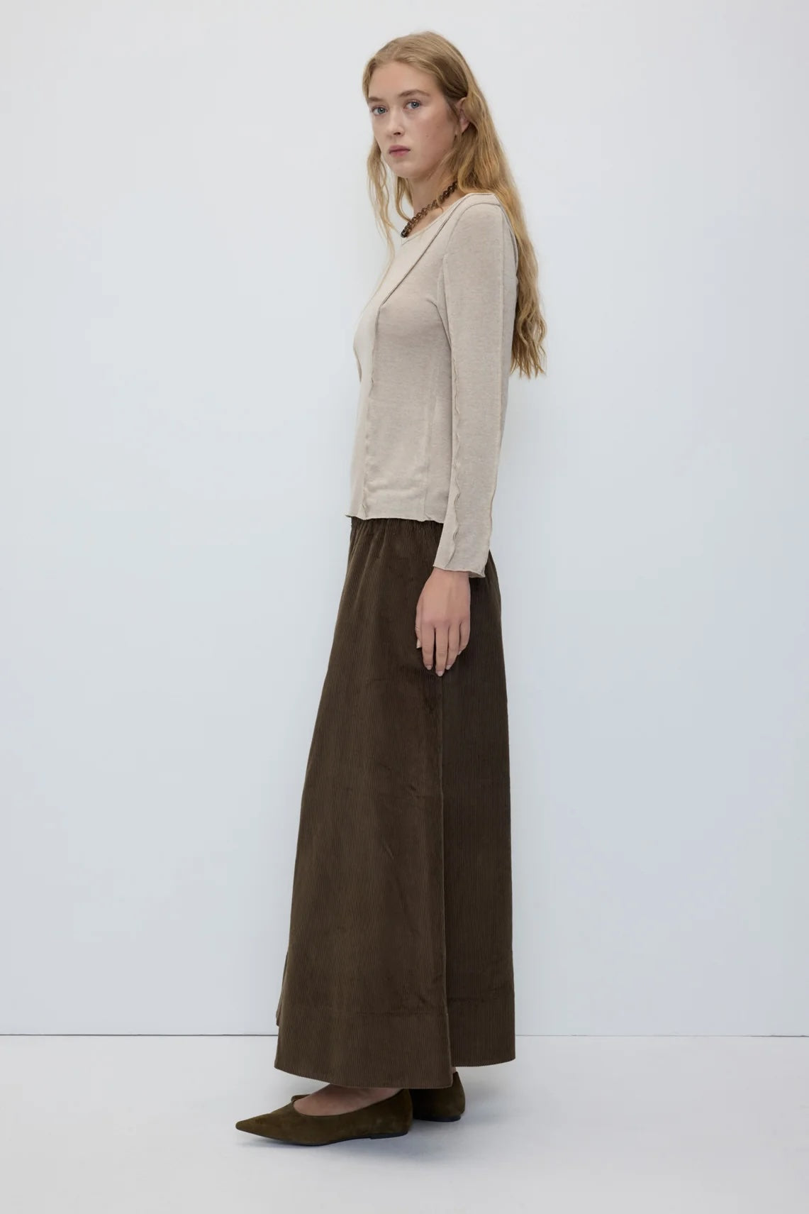 A woman stands in profile wearing a light beige long-sleeve knit top, a dark brown wide-leg skirt, and olive green flats against a plain white studio background.
