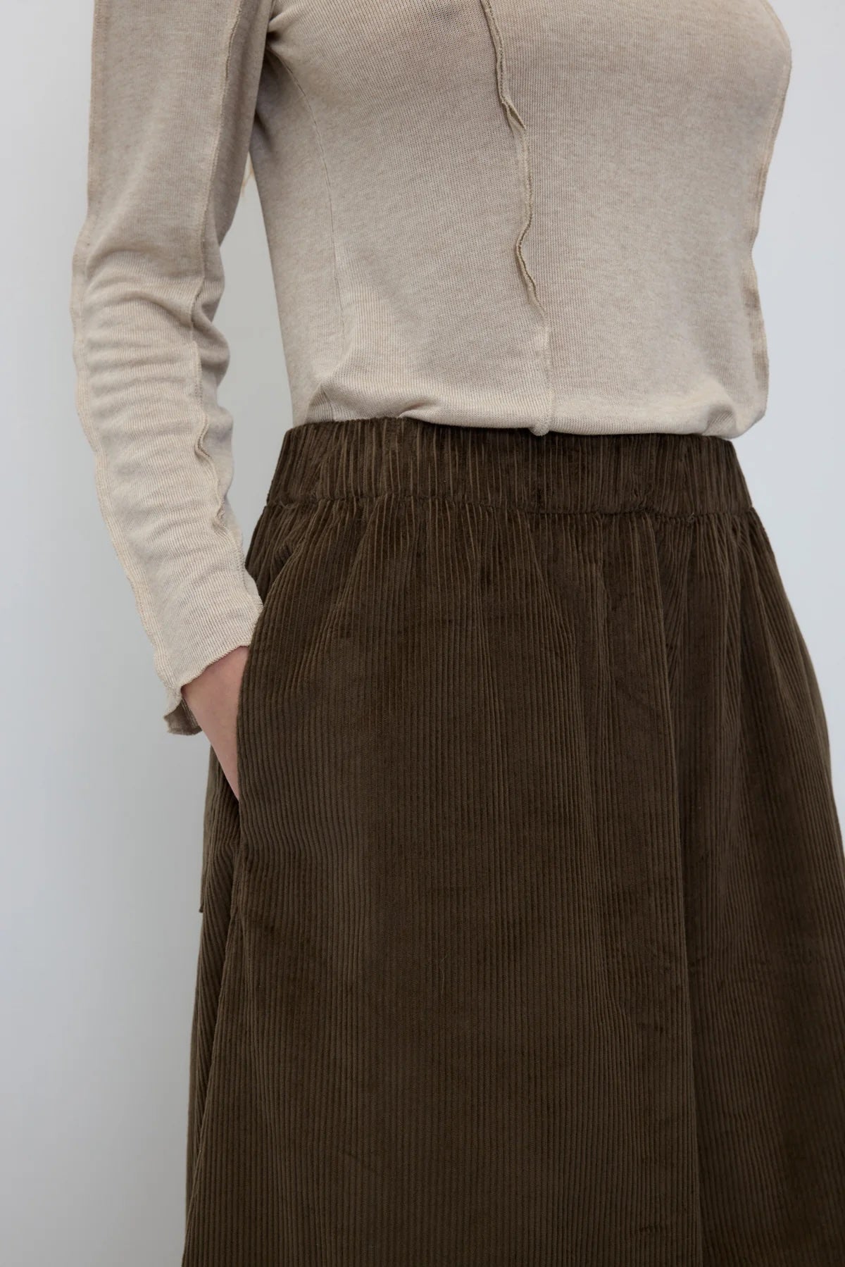 A close-up of a person wearing a beige long-sleeve knit top and a dark brown corduroy skirt with an elastic waistband and side pockets.