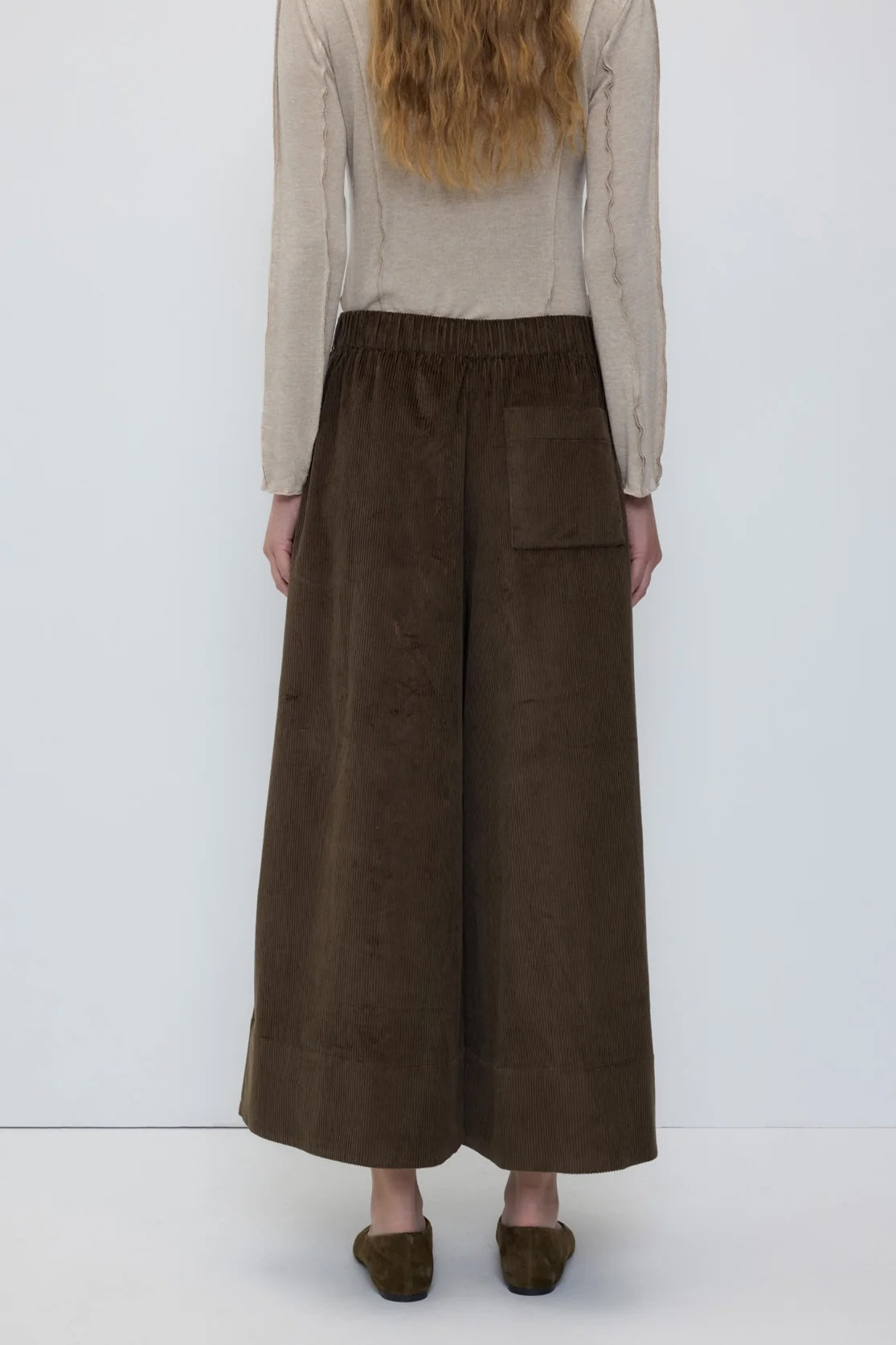 A model shown from the back wearing wide-leg brown corduroy trousers, a beige knit sweater, and brown slip-on shoes against a plain white studio background.