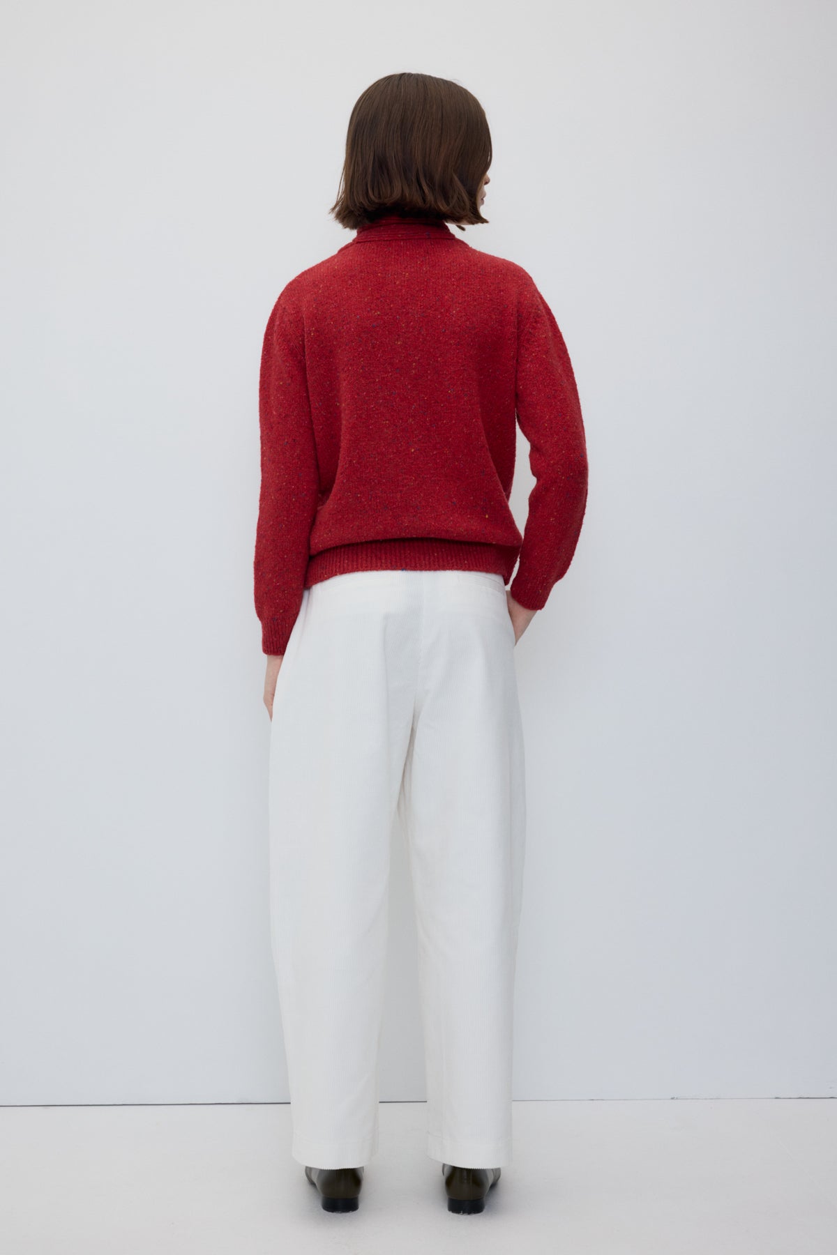 A woman stands in profile wearing a textured red cardigan, white wide-leg trousers, and dark brown shoes against a plain white studio background.