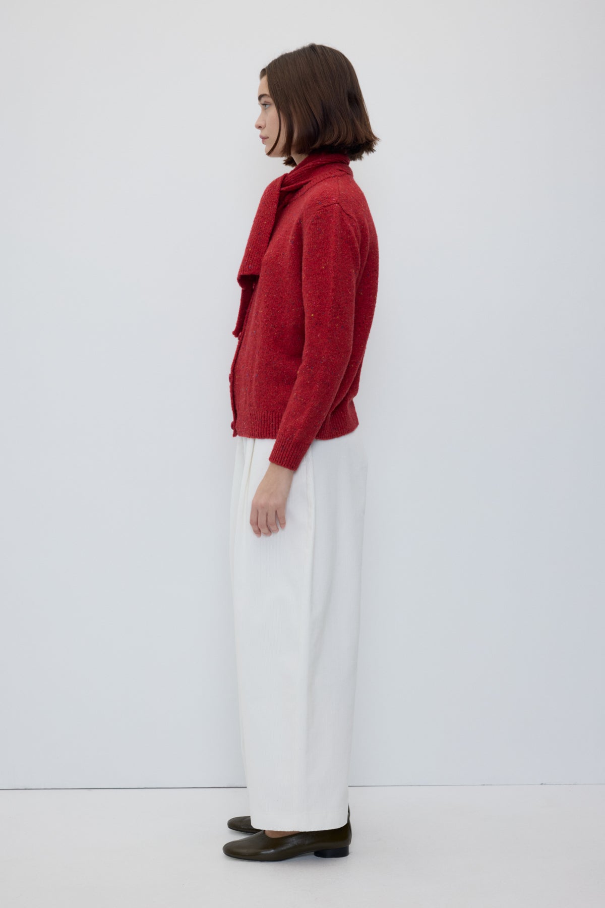 A woman stands in profile wearing a textured red cardigan, white wide-leg trousers, and dark brown shoes against a plain white studio background.