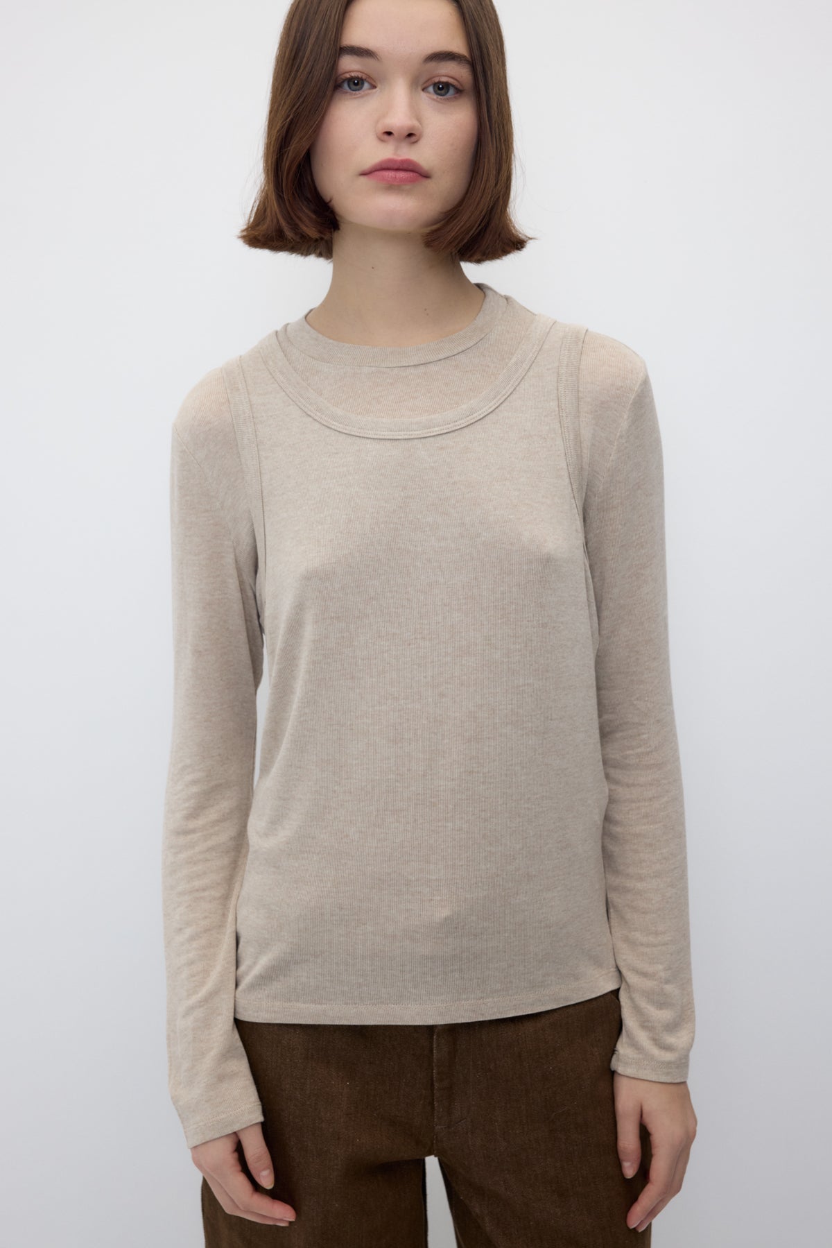 A woman with short brown hair wearing a light beige long-sleeve crewneck top and brown pants, standing against a plain light gray background.