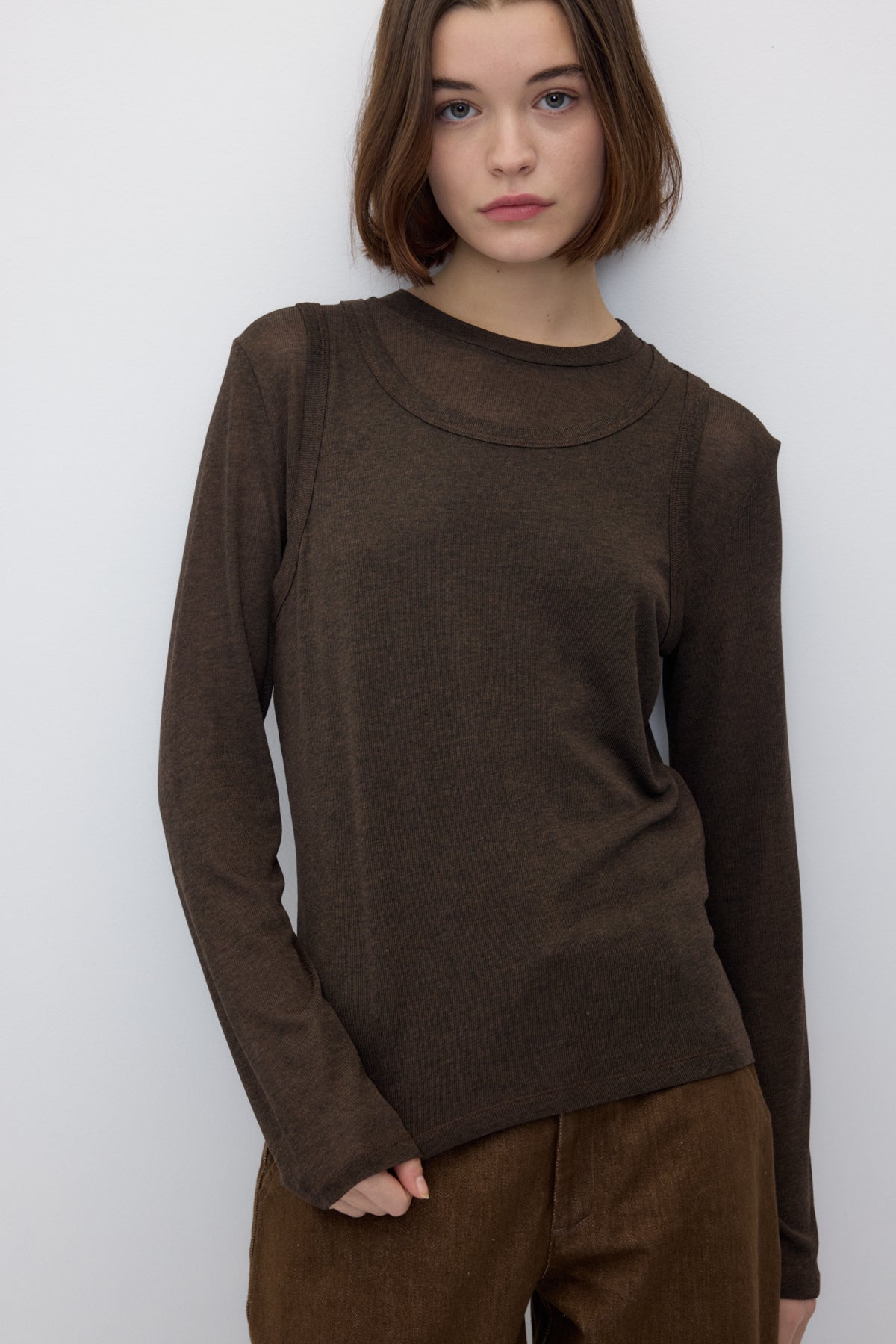 A model wearing a dark brown long-sleeve knit top with a wide mock neck and draped shoulder details, paired with brown corduroy pants, against a plain light gray background.