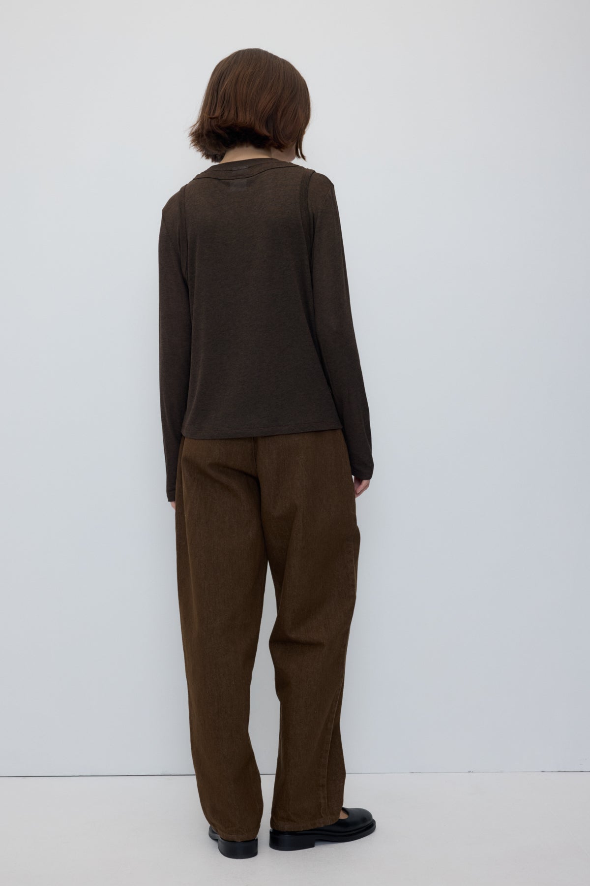 A model viewed from the back wearing a dark brown long-sleeve top and matching brown wide-leg trousers, standing against a plain white studio background.