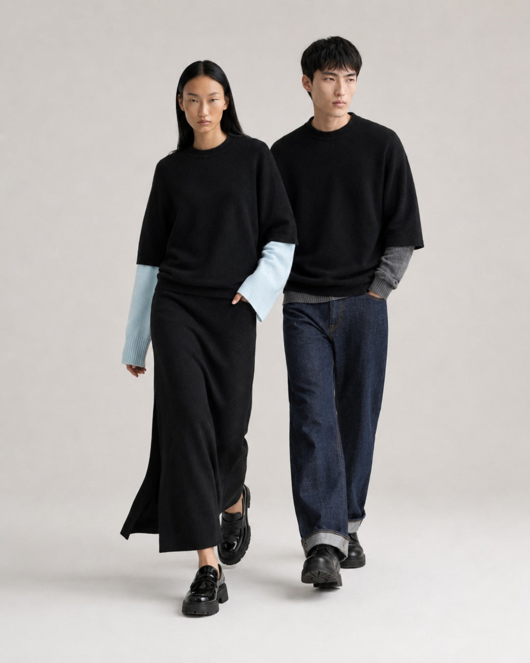 A man and a woman modeling minimalist black knit clothing against a light gray studio background. The woman wears a long-sleeve black top and skirt with light blue long sleeves, and the man wears a black crewneck sweater over a gray long-sleeve shirt with dark blue jeans.