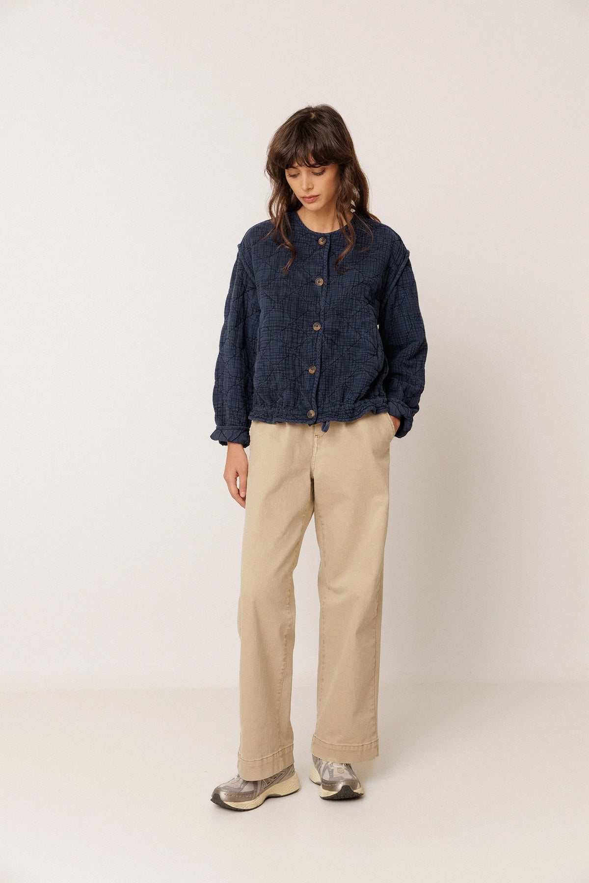 A model stands against a plain off-white studio background wearing a dark navy blue cropped button-up jacket, beige wide-leg trousers, and gray sneakers.
