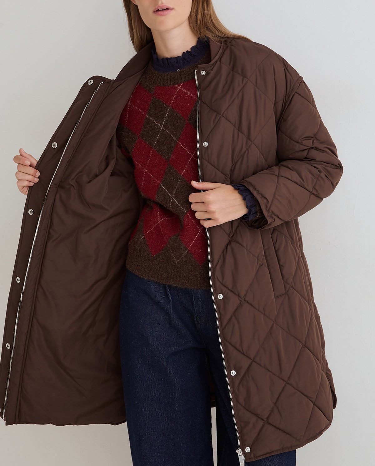 A woman wearing a brown quilted puffer jacket over a red and brown argyle sweater and blue jeans, shown against a plain light gray background.