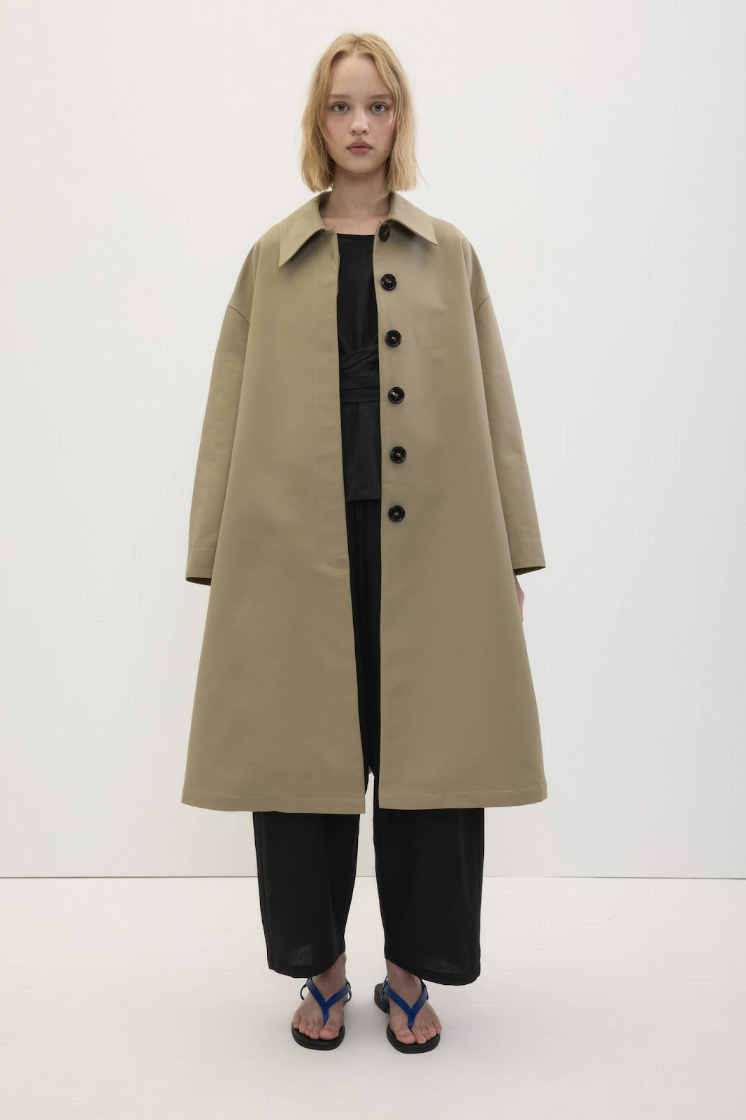 A model stands against a plain white studio background wearing a long, oversized beige trench coat over a black outfit, paired with black pants and blue flip-flops.