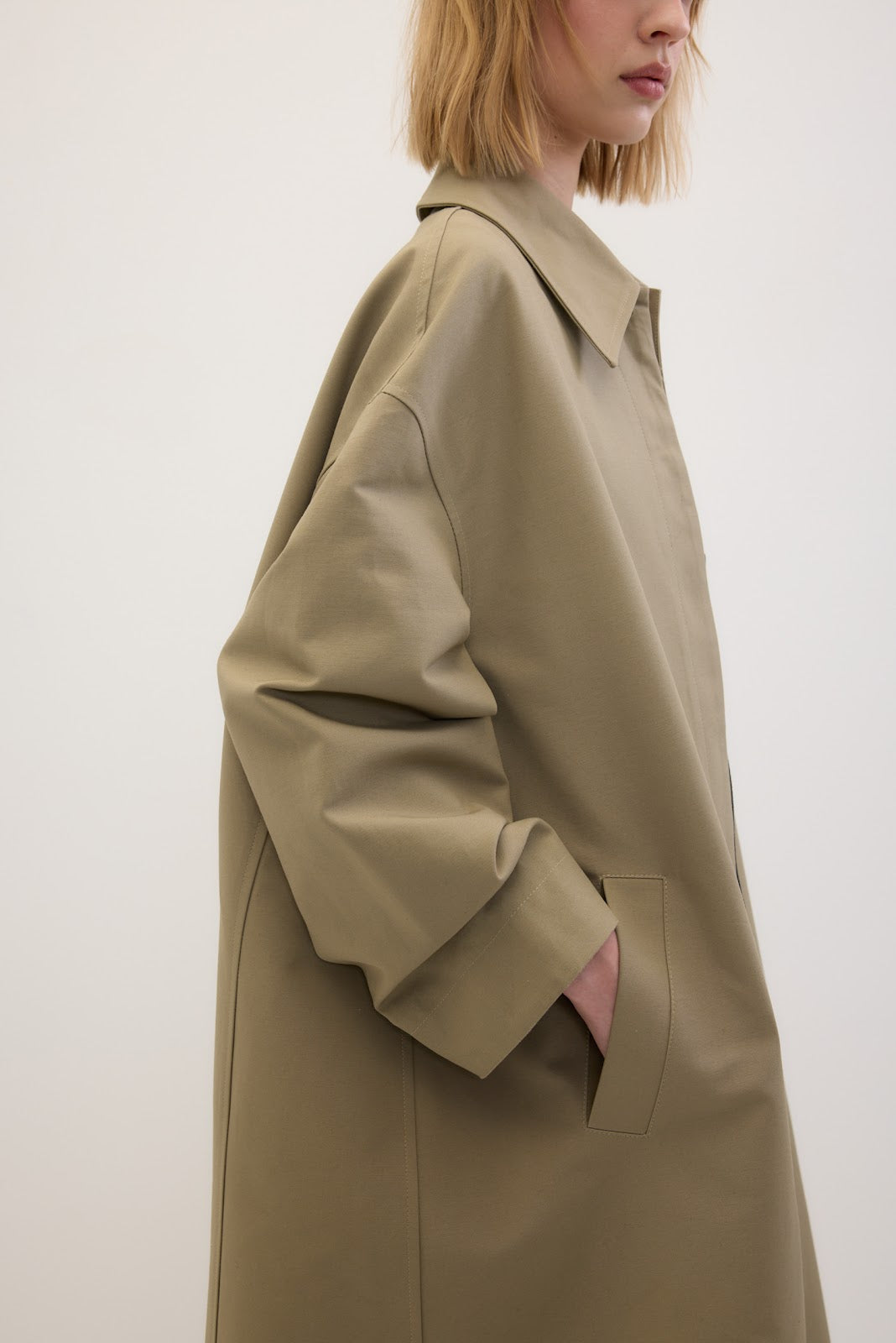 A side-profile view of a model wearing an oversized, light olive-beige trench coat against a plain off-white background.