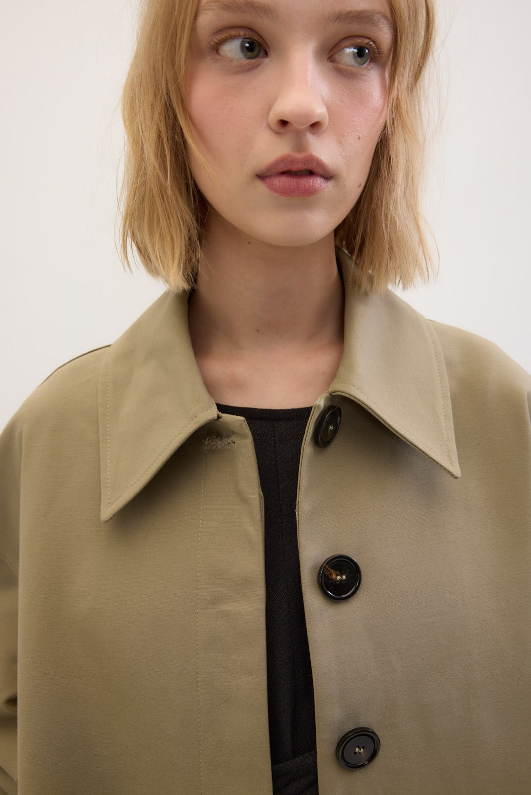 A close-up shot of a model wearing a light khaki trench coat over a black top, set against a plain off-white background.