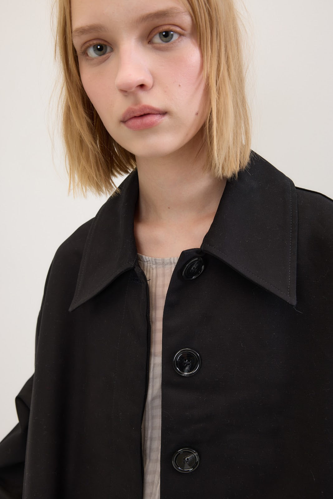 A close-up of a model wearing a black oversized button-up coat over a light beige ribbed top, set against a plain off-white background.