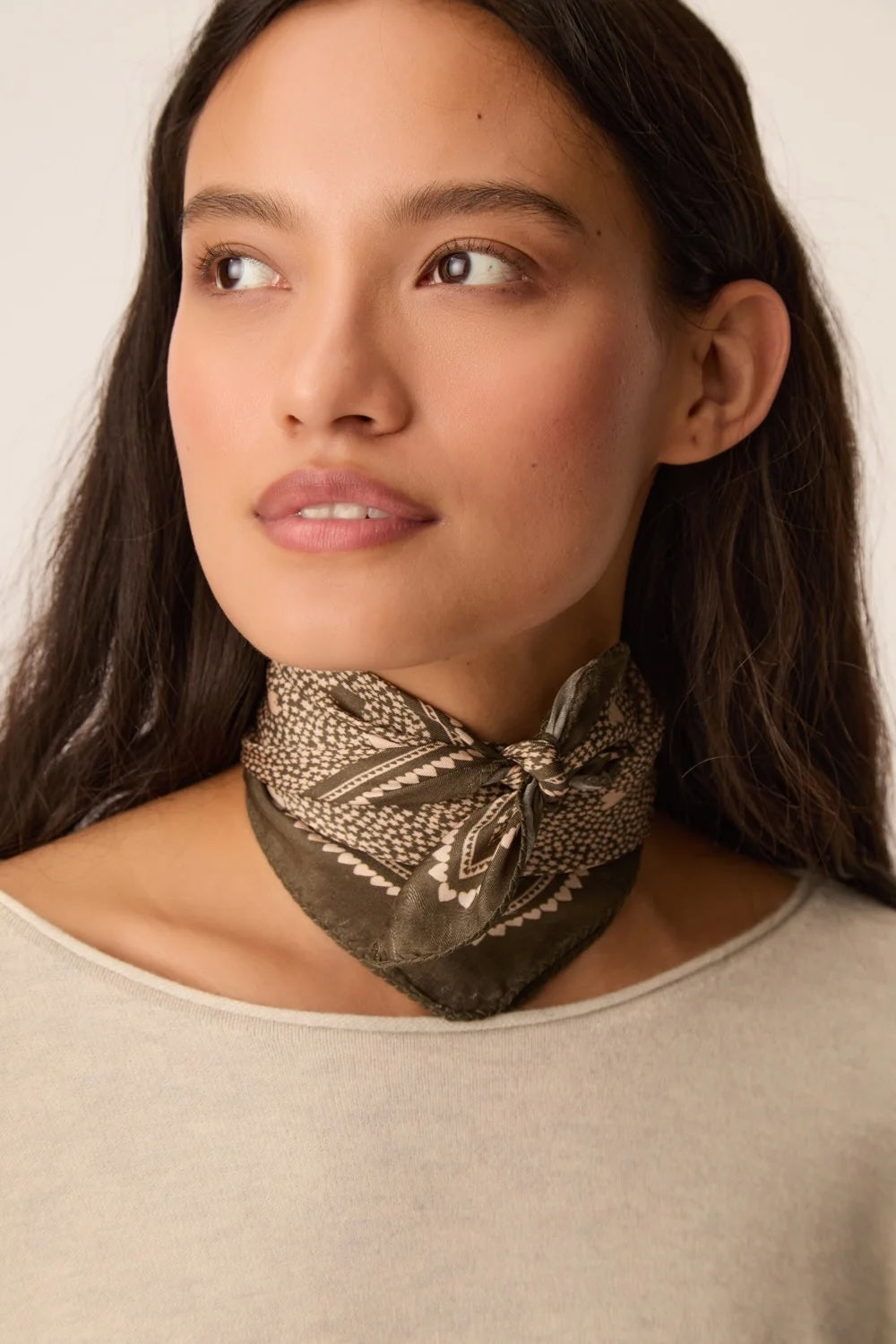 A woman wearing a dark olive green neck scarf with a beige geometric pattern, styled in a knot at the base of her neck.