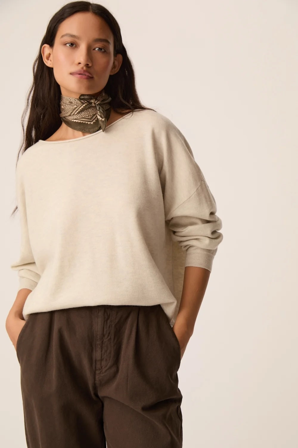 A woman modeling a cream-colored, wide-neck knit sweater with long sleeves, paired with dark brown trousers and a patterned neck scarf, set against a plain off-white background.