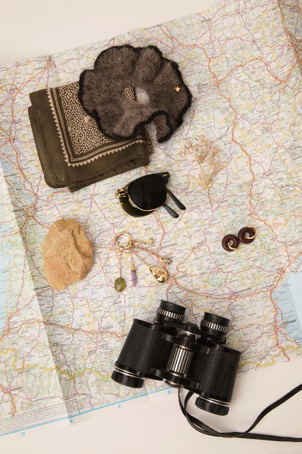 A styled flat-lay of various accessories arranged on an open road map, including black binoculars, a dark brown bandana, a black and brown patterned wallet, black sunglasses, gold-toned jewelry, and a small beige stone.