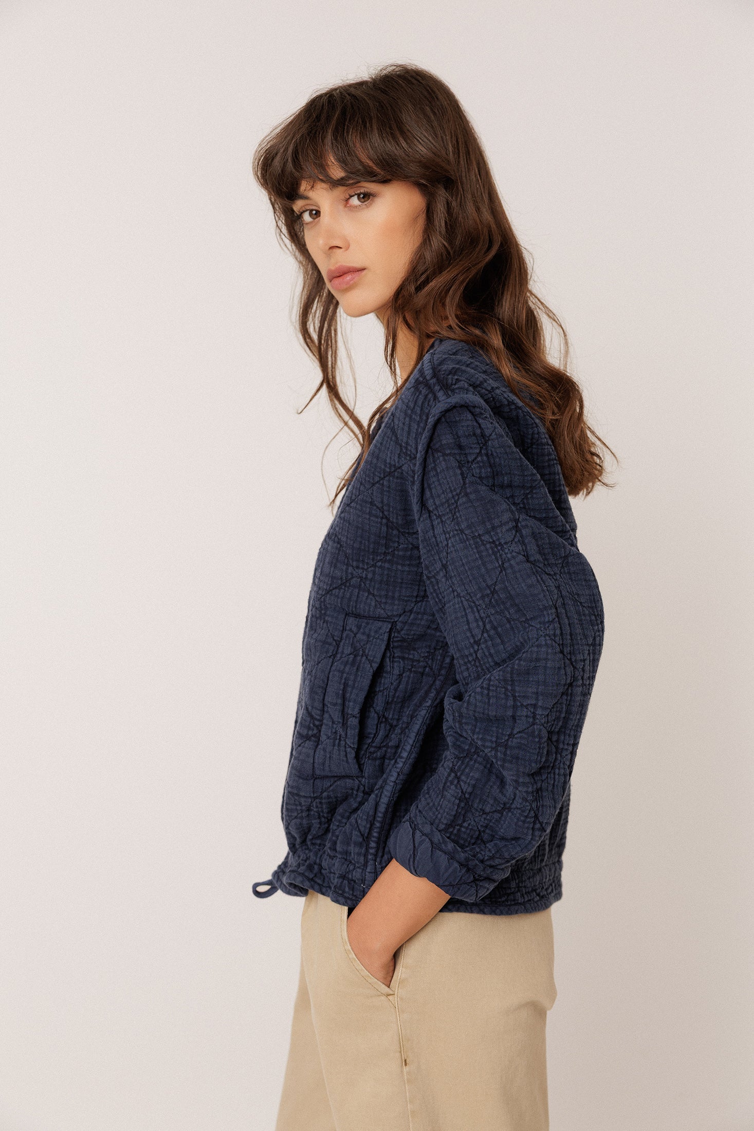 A woman wearing a textured navy blue cropped jacket with drawstrings and beige trousers, photographed against a plain light background.