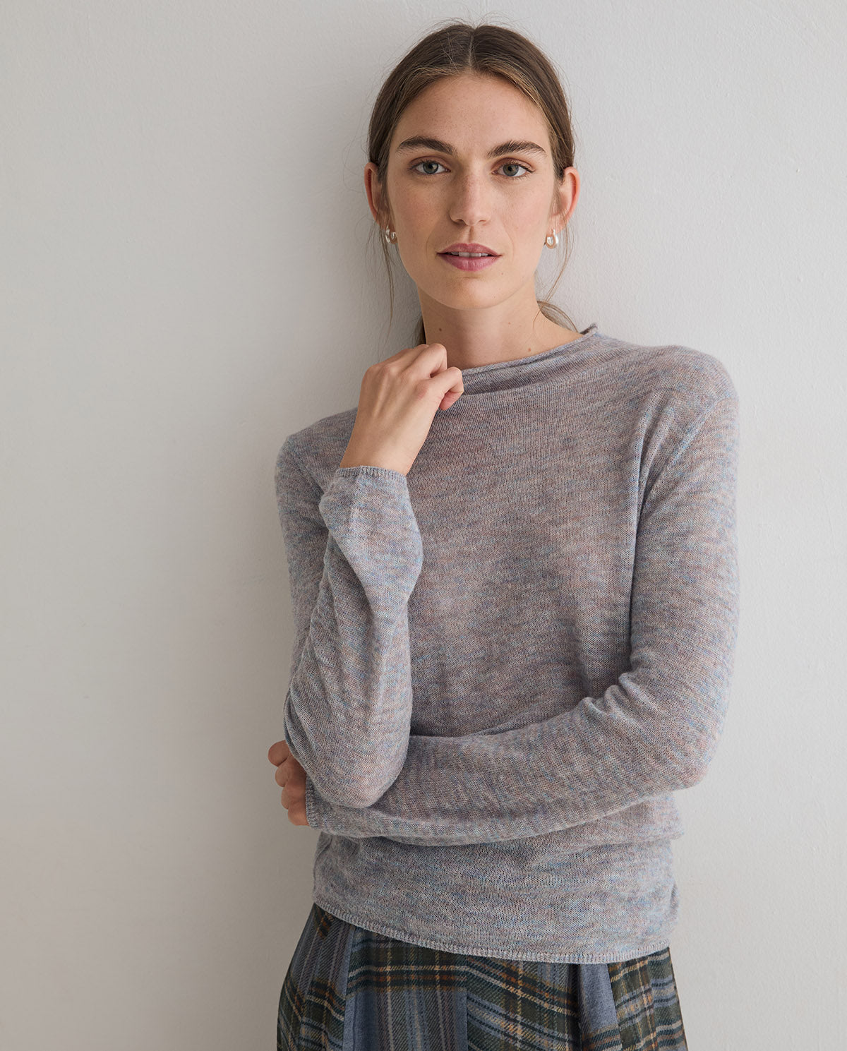 A woman wearing a light gray heathered long-sleeve knit sweater and a blue plaid skirt, posing against a plain light gray wall.