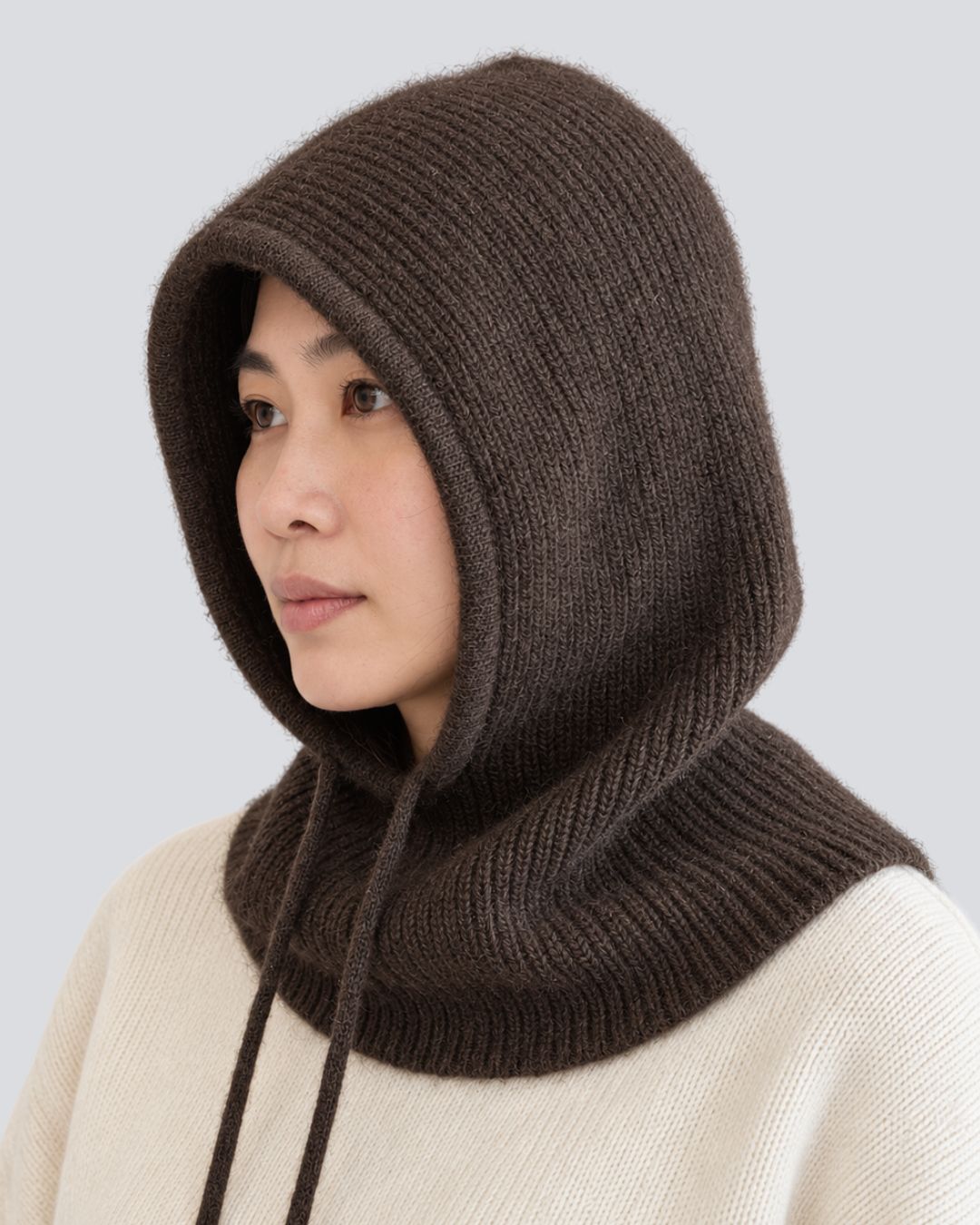 A woman wearing a dark brown ribbed knit hooded neck warmer over a cream-colored knit sweater, shown in a side profile against a light gray background.