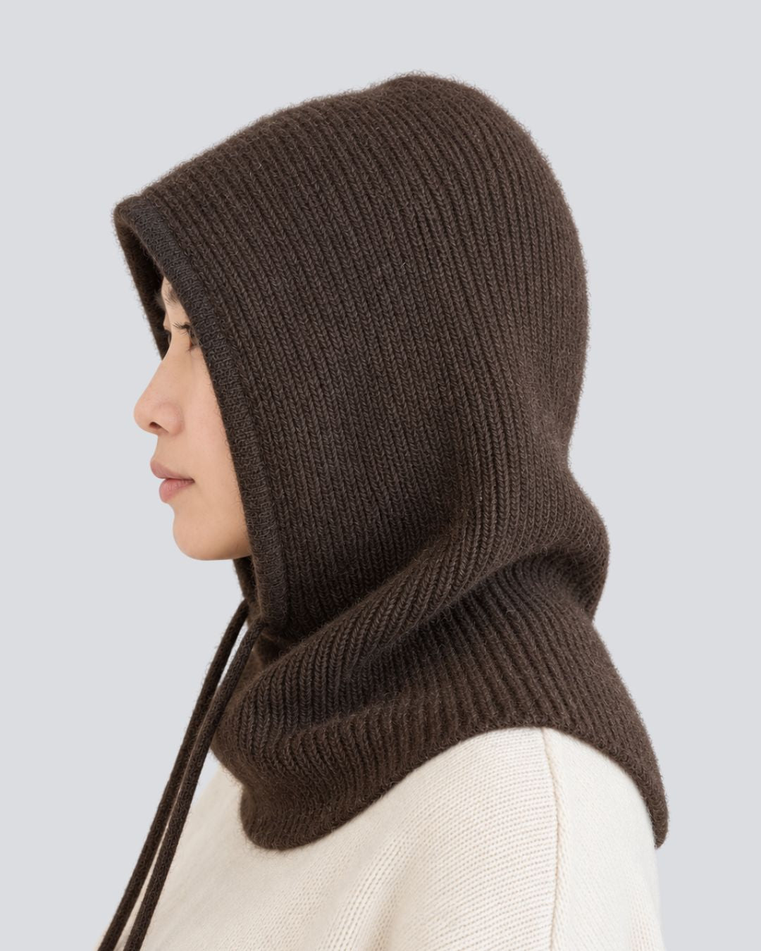 A side-profile view of a person wearing a dark brown ribbed knit hood with a matching neck gaiter and drawstrings over a cream-colored knit top, set against a solid light gray background.