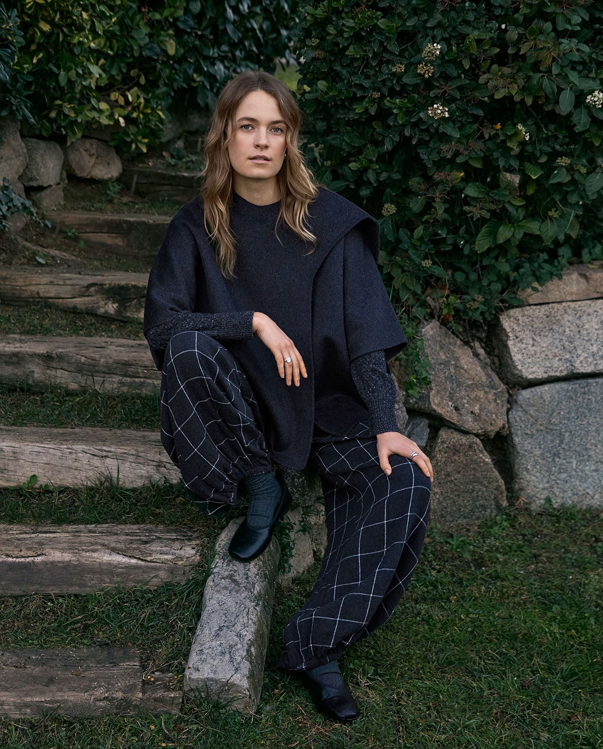 A woman sits on rustic wooden garden steps wearing a dark navy blue knit poncho-style top and matching navy plaid trousers, paired with black boots, set against a background of green foliage and stone.