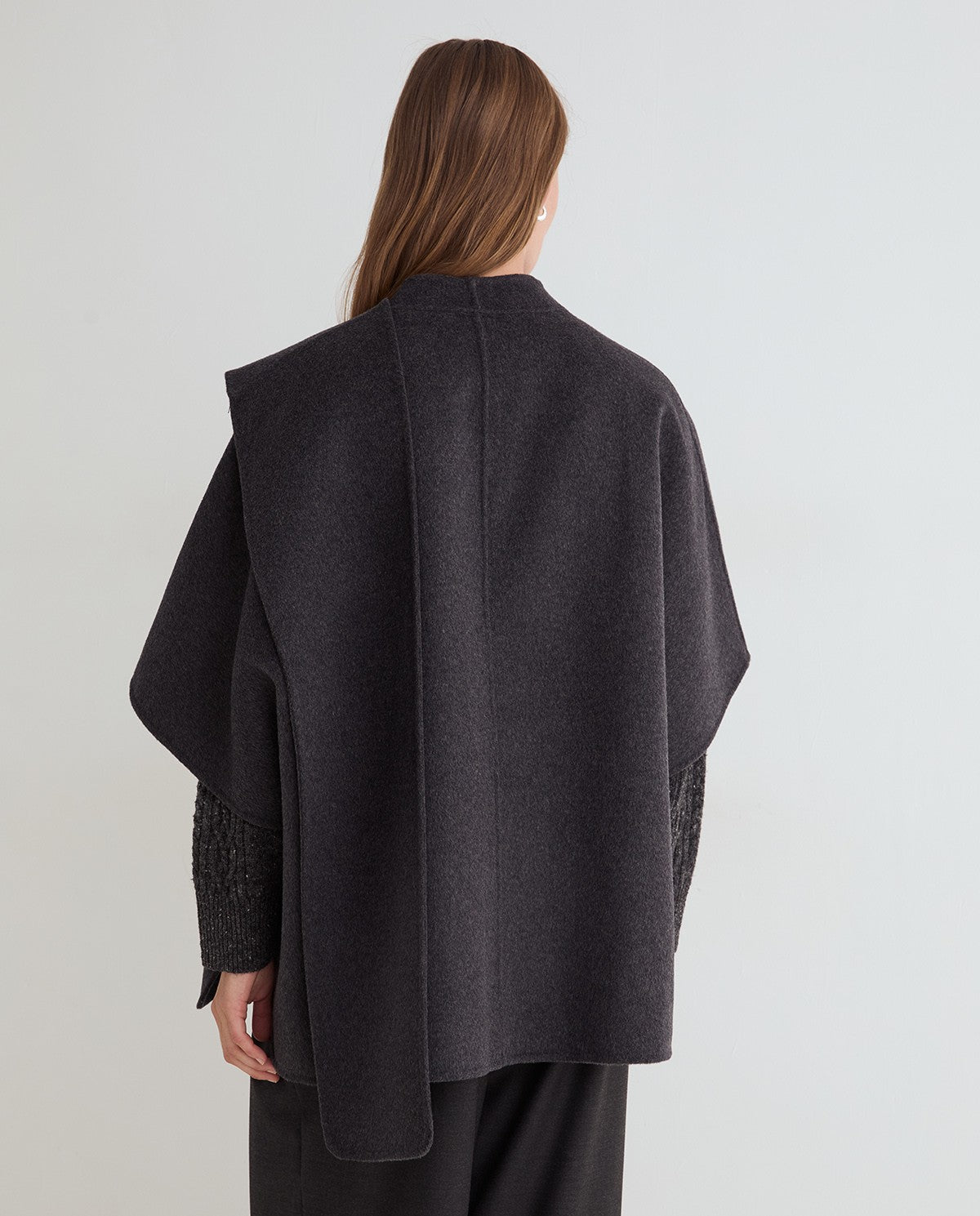 A woman shown from the back wearing a dark charcoal gray cape-style wrap jacket with textured knit sleeves, set against a plain light gray studio background.