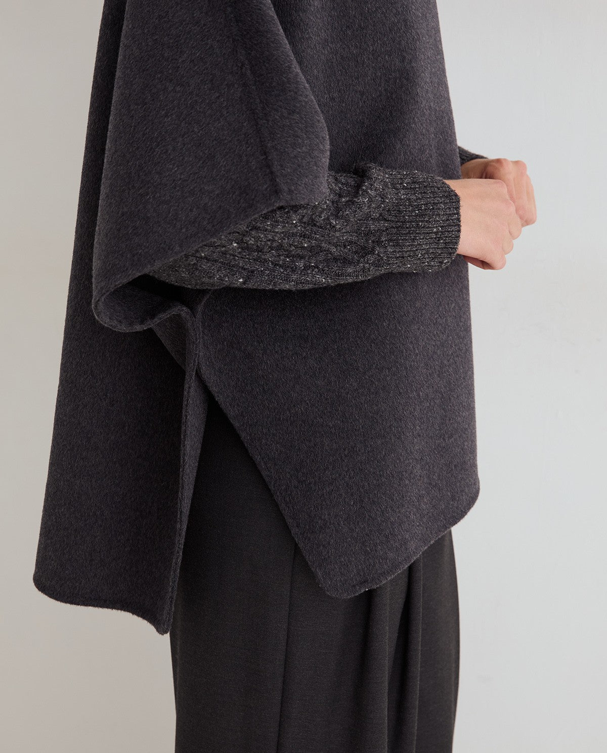 Close-up of a person wearing a dark charcoal gray draped poncho-style top over black wide-leg pants, showing the sleeve with a textured knit cuff and subtle glittery detailing.